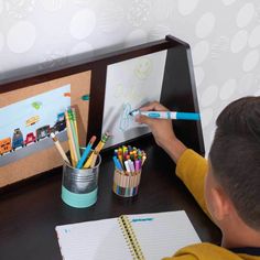 a young boy sitting at a desk with markers and pencils in front of him