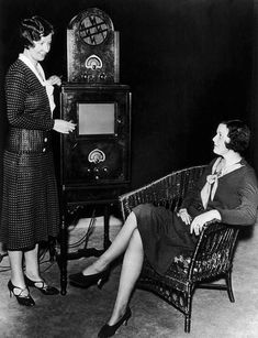 two women are talking to each other in front of an old fashioned radio and cabinet