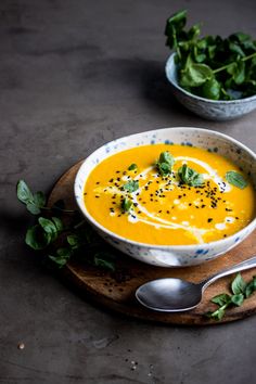 a bowl of carrot soup on a wooden board with two bowls of greens in the background