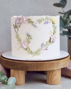 a white cake with flowers on it sitting on a wooden stand next to a potted plant