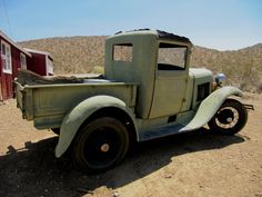 an old green truck is parked in the dirt near a red building and some hills