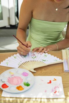 a woman in a green dress is painting with her hands and holding a fan while sitting at a table