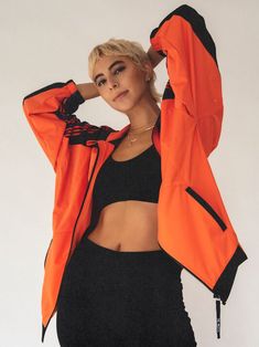 a woman wearing an orange jacket and black pants with her hands on her head, posing for the camera