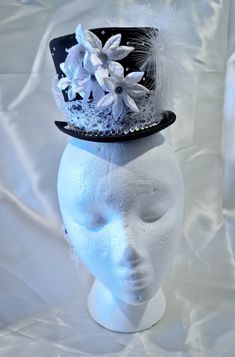 This hat is excellent for making a statement, its neautral colors allow you to build it into a focal point with any outfit. This satin miniature top hat can be worn with a headband or affixed to the wearers head via small loops inside the hat. Delicate white lace makes up the band, creating a stark contrast with the satin hat and forming a generous bow and waterfall tail in the back. The front of the band is decorated with a line of large rhinestones. Smaller rhinestones scatter thier glittery light around the rest of the hat. The front of the hat features small white velvet poinsettia flowers, with small gear and cog centers. The side features white feathers with a black and white cameo. Whimsical Mini Hats For Parties, Handmade High Crown Top Hat For Party, Handmade Fitted High Crown Mini Hats, Adjustable Short Brim Top Hat For Costume, Handmade Fitted Mini Hat With High Crown, Elegant Tall Crown Adjustable Hat, Brimmed Party Costume Hats And Headpieces, Whimsical Adjustable Costume Accessories, Adjustable Tall Crown Costume Hats For Party