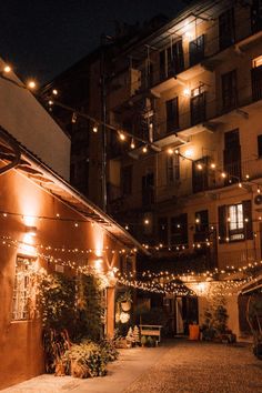 an alley way with lights strung from the buildings and potted plants on either side