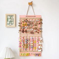 a pink wall hanging with lots of bracelets and hair clips on it next to a lamp