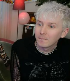 a woman with short white hair wearing a black shirt and silver necklace sitting in front of a christmas tree