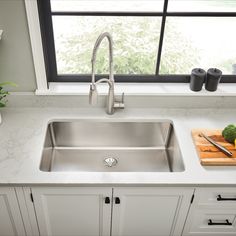 a stainless steel kitchen sink in front of a window with cutting board and utensils on the counter