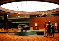 three people standing in front of a fountain with lights on the ceiling and two lamps hanging from the ceiling