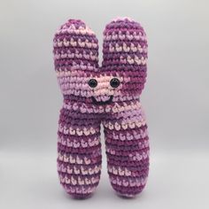 a crocheted purple and white teddy bear with black eyes sitting in front of a gray background