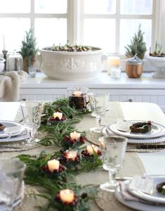 the table is set for christmas dinner with candles and greenery on it, along with other place settings