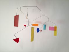 a mobile made out of colored paper hanging from a white wall next to a red, yellow, blue and pink piece of art