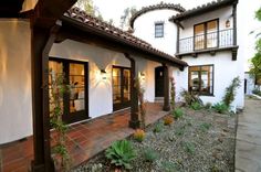 a white house with black doors and brown trim on the front door is surrounded by greenery