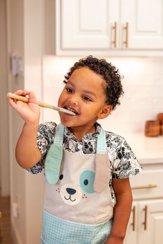 a little boy brushing his teeth in the kitchen with an apron on and a toothbrush in his mouth