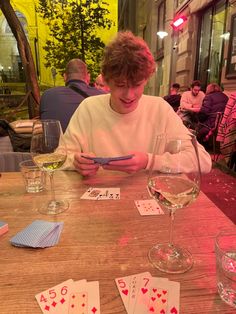 a young man sitting at a table with playing cards and wine glasses in front of him