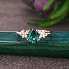 an emerald and diamond ring sitting on top of a green piece of wood with flowers in the background