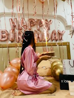 a woman sitting on the floor holding a cake with lit candles in her hand and balloons behind her