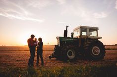 two people standing next to a tractor in a field with the sun setting behind them