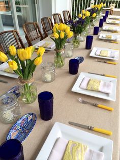 a long table with yellow flowers in vases and plates on it, along with other place settings