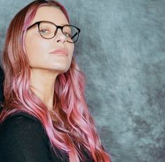 a woman with pink hair and glasses looking off into the distance while standing in front of a gray wall