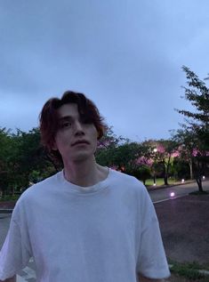 a man with red hair standing in front of a parking lot at dusk, wearing a white t - shirt