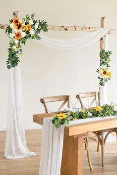 a wooden table topped with flowers and greenery next to a white drape covered wall