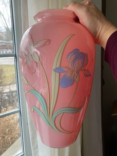 a pink vase with purple flowers painted on it is being held up by a woman's hand