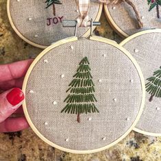 three hand embroidered christmas ornaments with trees on them, one being held by a woman's hand