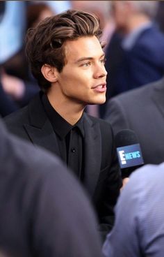 a young man in a black suit talking to the camera while holding a microphone and looking off into the distance