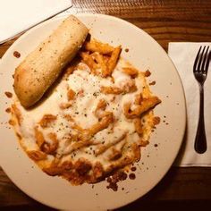 a white plate topped with pasta covered in cheese and sauce next to a roll on top of a wooden table