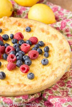 a pie with blueberries and raspberries on top sits on a table next to lemons