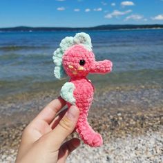 a hand holding a small pink crocheted stuffed animal in front of the ocean