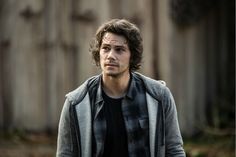 a man with curly hair wearing a gray jacket and black t - shirt standing in front of a wooden fence