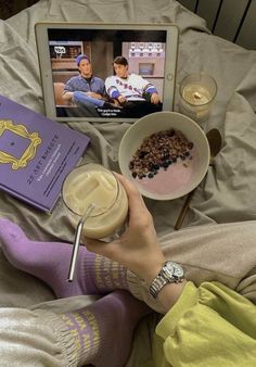 a person laying on a bed with food and a book in front of them while watching tv