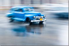 an old blue truck is driving down the street in the rain with other cars behind it