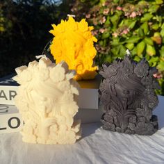 three different colored sculptures sitting on top of a white tablecloth covered table next to trees