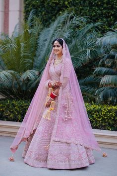 a woman in a pink lehenga and veil posing for the camera with palm trees behind her