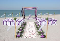 an outdoor wedding setup on the beach with purple and white flowers, ribbons and chairs