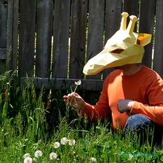 a person sitting in the grass with a paper mask on their head and a dandelion