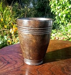 a metal cup sitting on top of a wooden table in front of some bushes and trees