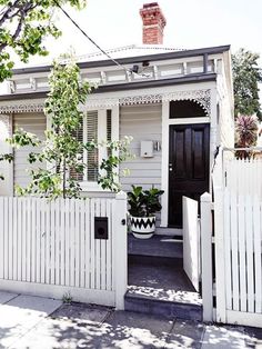 a small white house with a black door and window on the front porch, next to a picket fence