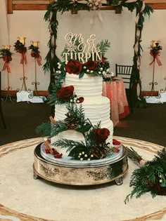 a white wedding cake with red roses and greenery on top sitting on a table