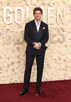 a man in a tuxedo standing on a red carpet with flowers behind him