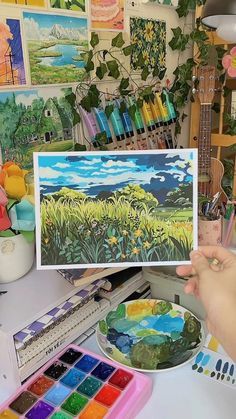 a person holding up a painting in front of a table with paints and watercolors