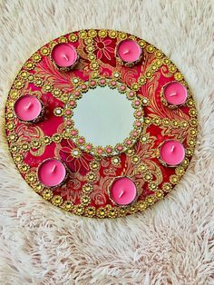 a red and gold circular mirror with candles on it sitting on a fluffy white surface