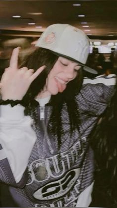 a woman with long hair wearing a baseball cap and jacket, making the peace sign