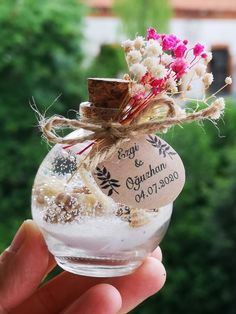 a hand holding a small glass jar filled with flowers and sand in front of trees