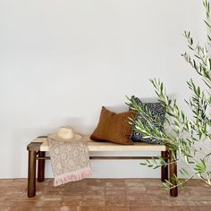 a wooden bench with pillows and a hat on it next to a plant in front of a white wall