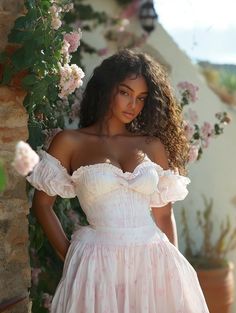 a woman in a white dress standing next to a flower covered wall with her hands on her hips