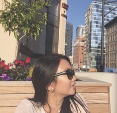 a woman sitting on top of a wooden bench in front of tall buildings and flowers